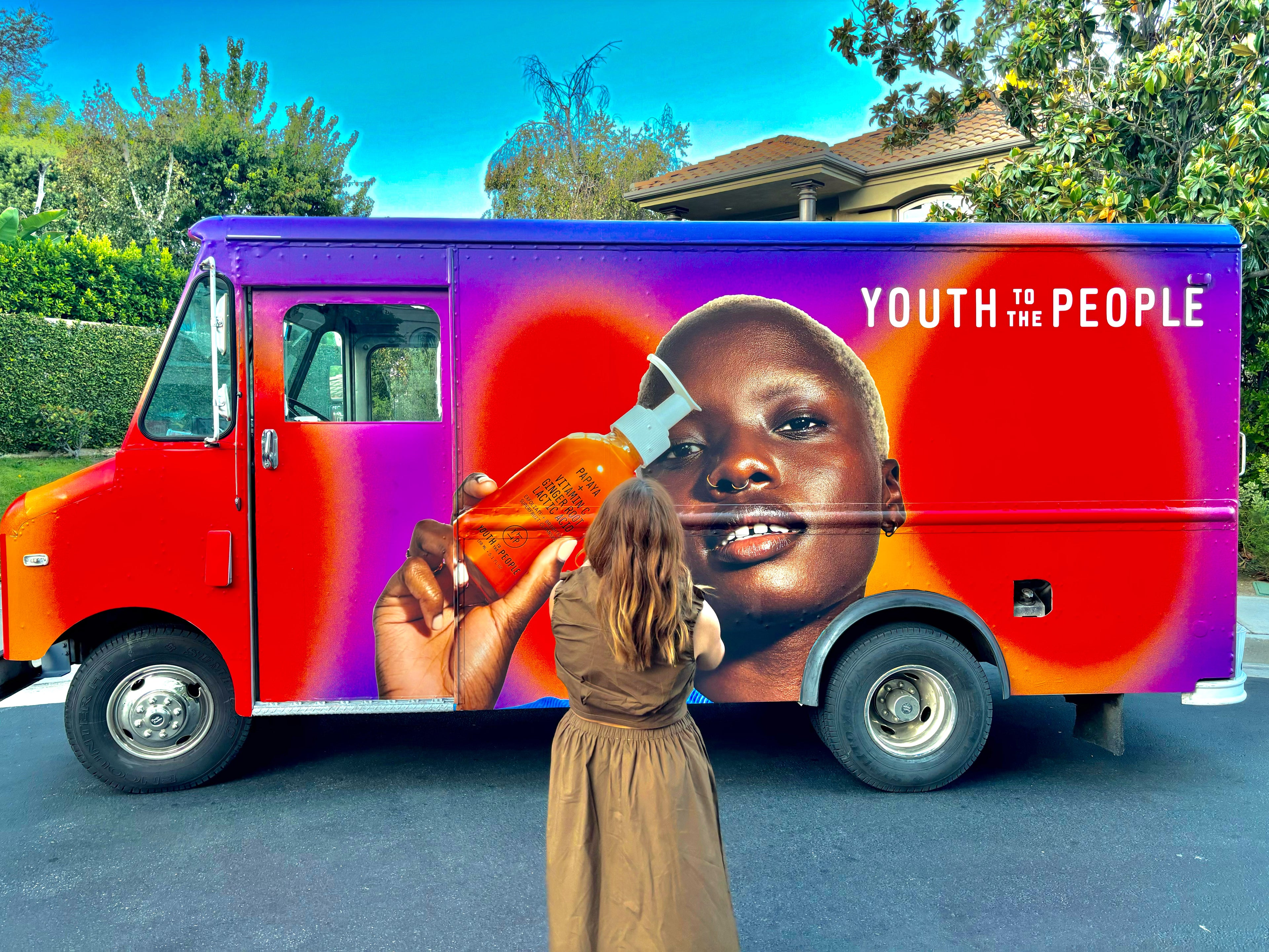 Colorful Sample truck with a large graphic of a models face advertising for a cosmetics brand, with a person standing in front taking a picture.